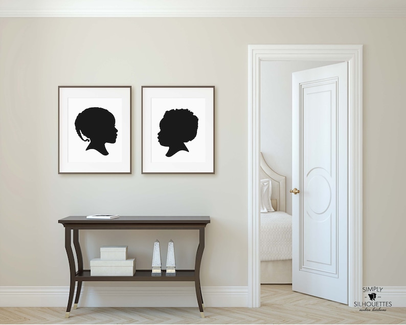 May include: Two black and white silhouette portraits of children framed in simple black frames, hanging on a white wall above a dark brown console table with a white box and two silver candle holders on the top shelf.