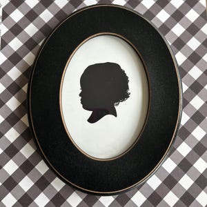 May include: Oval black picture frame with a black silhouette of a person's profile. The silhouette is centered on a white background. The frame is set against a black and white checkered pattern.