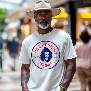 May include: A white t-shirt with a circular graphic featuring a blue silhouette and the text "WHO IS THE MASTER?" and "SHO'NUFF". The shirt is worn with a light brown hat and khaki pants. The image is taken outdoors.