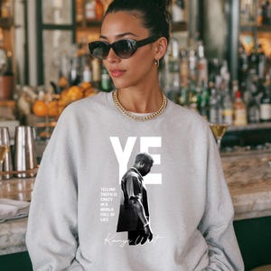 May include: A light gray sweatshirt featuring a black and white image of a person with the word "YE" in white block letters. The text "TELLING TRUTH IS CRAZY IN A WORLD FULL OF LIES" and "Kanye West" are also printed on the sweatshirt.