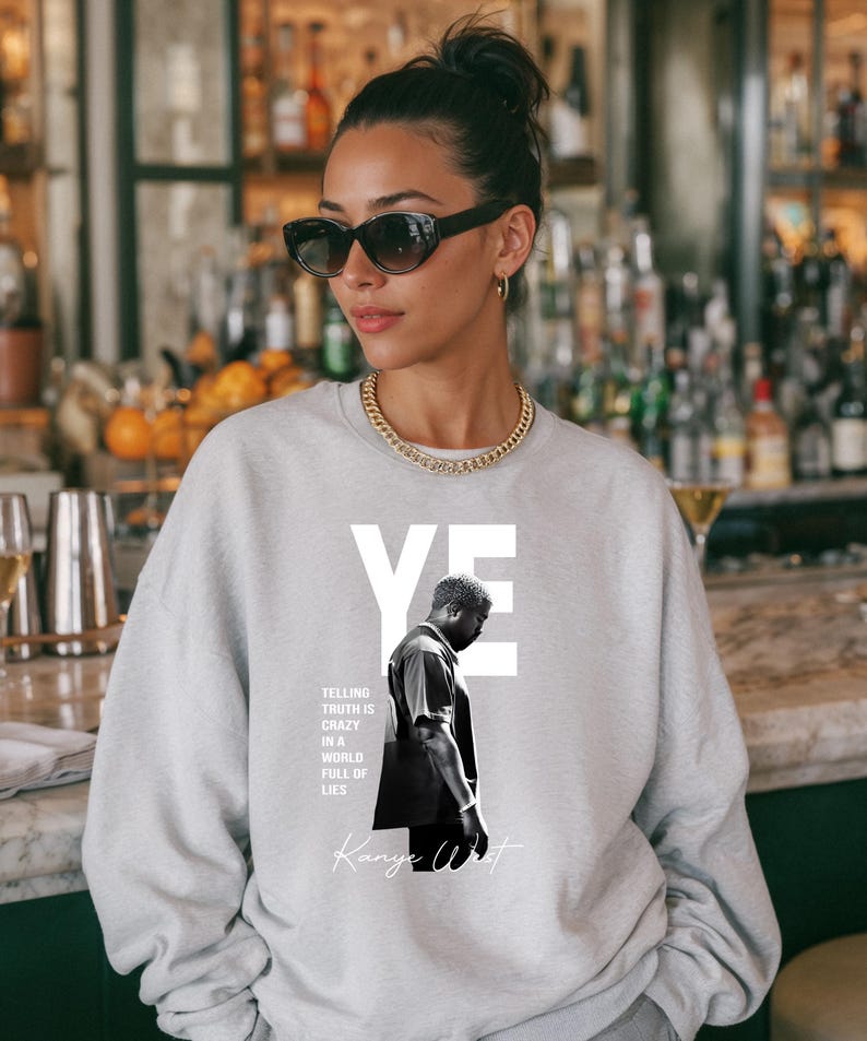 May include: A light gray sweatshirt featuring a black and white image of a person with the word "YE" in white block letters. The text "TELLING TRUTH IS CRAZY IN A WORLD FULL OF LIES" and "Kanye West" are also printed on the sweatshirt.