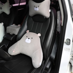May include: A set of three gray car seat cushions with a bear face design. The cushions are made of soft, plush fabric and are designed to provide comfort and support while driving. The cushions are placed in a car seat, with one on the headrest, one on the back, and one on the armrest.
