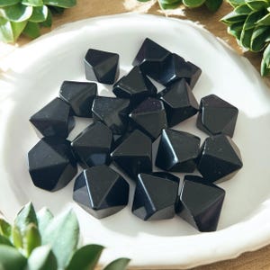 Op de afbeelding: Een verzameling gepolijste, gefacetteerde zwarte obsidiaanstenen gerangschikt op een wit bord. De stenen hebben een geometrische vorm en reflecteren licht, waardoor een glanzend uiterlijk ontstaat. Groene planten omringen het bord.