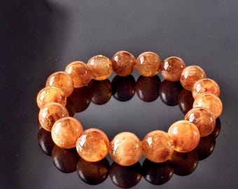May include: A circular bracelet composed of polished, translucent orange and amber-toned spherical beads. The beads exhibit internal variations in color density and texture, creating a marbled appearance throughout the round gemstone structure.