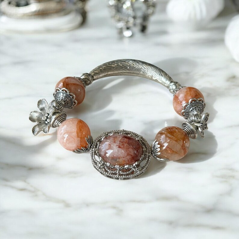 May include: A silver-toned bracelet featuring large, faceted, peach-colored beads and a central oval stone. The bracelet has decorative floral and feather-like accents.