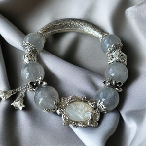 May include: A silver bracelet with gray gemstone beads and a decorative feather-shaped clasp. The bracelet features a cameo pendant and floral accents. The beads are approximately 0.75 inches in diameter.