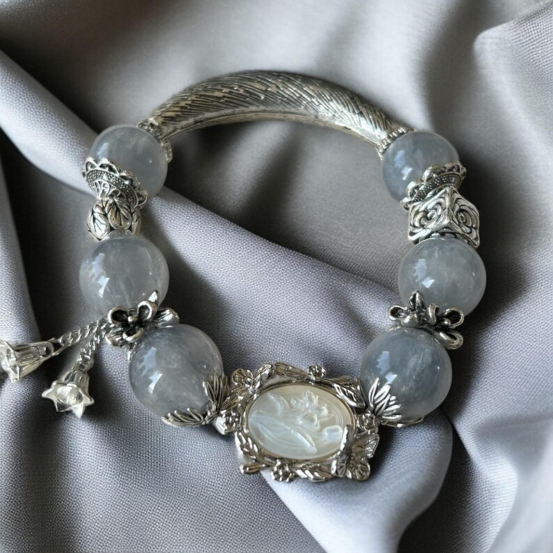 May include: A silver bracelet with gray gemstone beads and a decorative feather-shaped clasp. The bracelet features a cameo pendant and floral accents. The beads are approximately 0.75 inches in diameter.