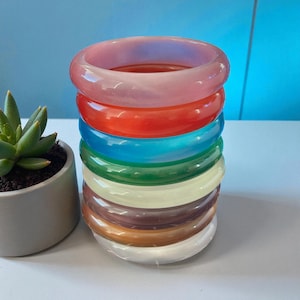 Pode incluir: Uma pilha de oito pulseiras translúcidas coloridas em tons de rosa, vermelho, azul, verde, amarelo, castanho e branco. As pulseiras estão empilhadas numa superfície branca, com uma pequena planta suculenta em vaso à esquerda.