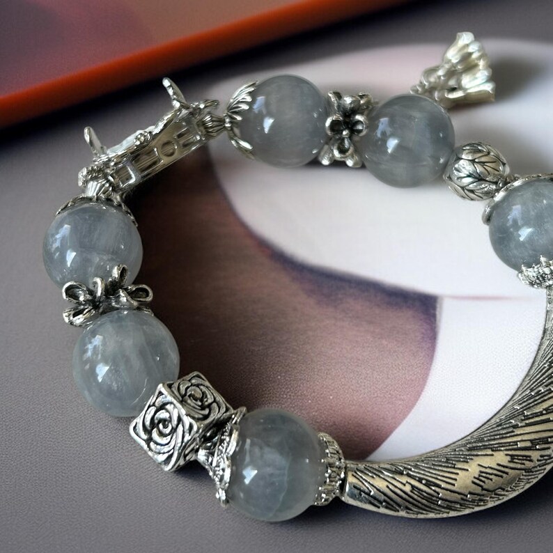 May include: A silver bracelet featuring large, round, gray beads. The bracelet has ornate silver accents, including floral and geometric designs. The clasp is shaped like a dragon's head. The bracelet is a piece of jewelry.