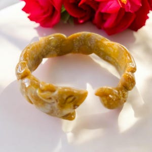 May include: A carved jade bangle bracelet with a light brown and white color. The bracelet is intricately carved with a floral design.