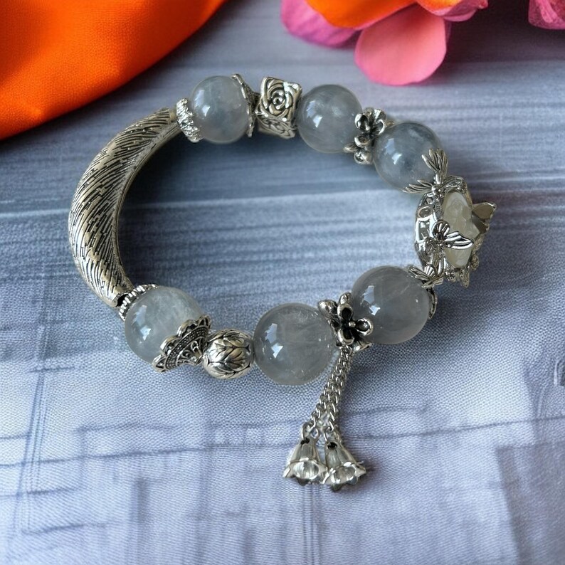 May include: A silver bracelet featuring large, round, gray beads interspersed with smaller decorative silver elements. The bracelet has a textured, curved silver piece and a dangling charm with bell-shaped accents. The bracelet is displayed on a patterned surface.
