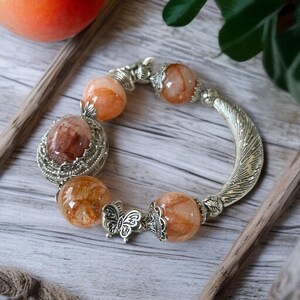 May include: A silver bracelet with orange and brown gemstone beads. The bracelet features a decorative butterfly clasp and a large oval-shaped center stone. The beads are round and have a translucent quality. The bracelet is displayed on a wooden surface.