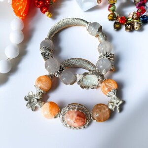 May include: Two bracelets with silver-toned metal accents and gemstone beads. One bracelet features orange and clear beads, while the other has clear beads. The bracelets are adorned with floral and oval-shaped embellishments.