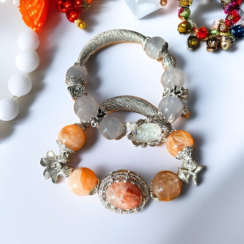 May include: Two bracelets with silver-toned metal accents and gemstone beads. One bracelet features orange and clear beads, while the other has clear beads. The bracelets are adorned with floral and oval-shaped embellishments.