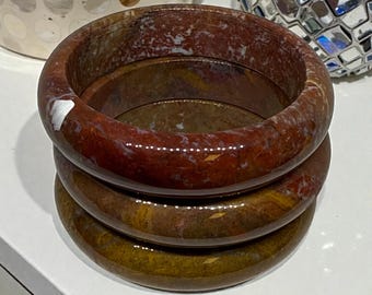 May include: Three stacked, polished stone bangles. The bangles are a mix of red, brown, and tan hues, with a marbled appearance. The smooth, rounded design of the bracelets suggests they are meant to be worn as jewelry.