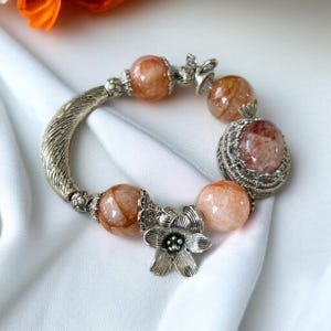 May include: A silver bracelet featuring large, round, peach-colored beads with reddish inclusions. The bracelet has a curved, textured silver band and is accented with silver floral and leaf-shaped embellishments. The bracelet is displayed on a white surface.