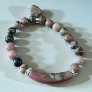 Peut inclure: Un bracelet de perles avec des perles de pierres précieuses roses, marron et noires. Le bracelet comprend une pièce maîtresse incurvée en pierre précieuse rose avec des accents argentés et un charm en forme de cœur. Les perles mesurent environ 1,3 cm de diamètre.