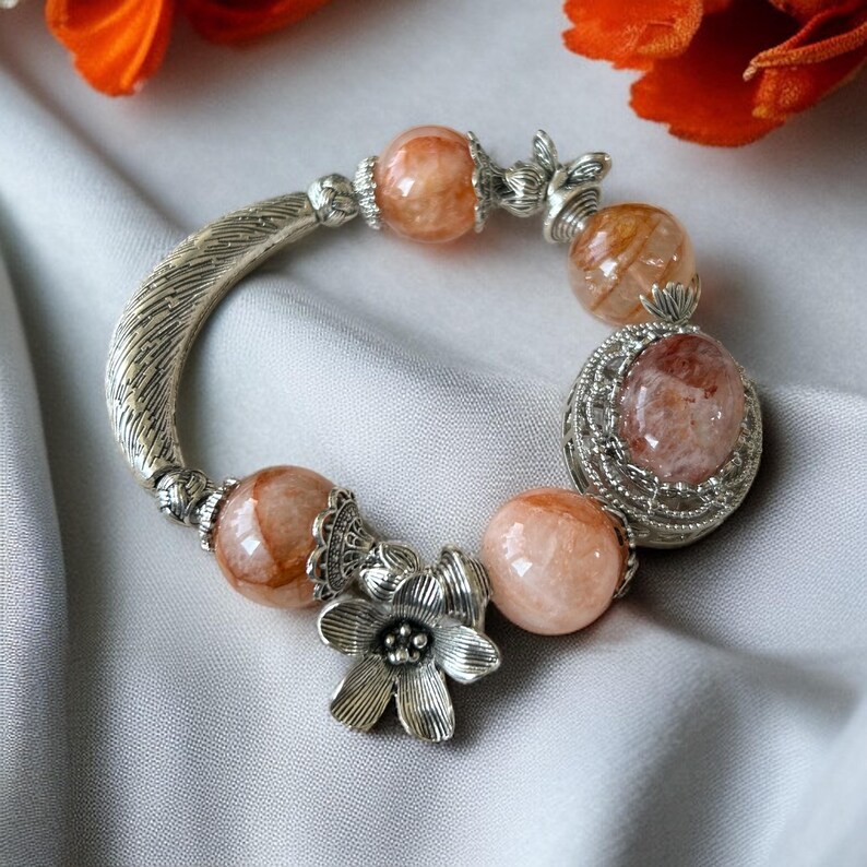 May include: A silver bracelet featuring peach-colored, round gemstone beads and decorative silver accents. The bracelet includes a flower charm and a curved, feather-like design. The beads are approximately 1 cm in diameter.