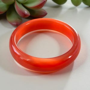 May include: A vibrant orange bangle bracelet. The smooth, polished surface reflects light, showcasing a gradient of color from a deep red-orange to a lighter, almost translucent hue. The bracelet is a circular shape, perfect for everyday wear.