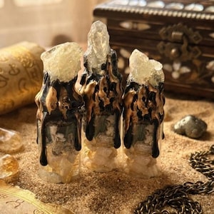 May include: Three clear glass vials filled with small light-colored stones, topped with jagged translucent crystals. The exterior of each vial is coated in dripping black and metallic gold paint that flows down from the crystal tops.