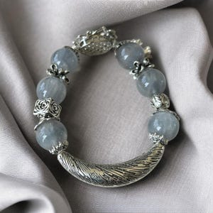 May include: A bracelet featuring large, round, pale blue beads interspersed with ornate silver-toned metal accents. The bracelet has a curved, textured silver clasp and decorative elements. The beads appear to be semi-translucent, and the bracelet is set against a neutral background.