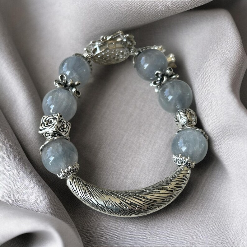 May include: A bracelet featuring large, round, pale blue beads interspersed with ornate silver-toned metal accents. The bracelet has a curved, textured silver clasp and decorative elements. The beads appear to be semi-translucent, and the bracelet is set against a neutral background.