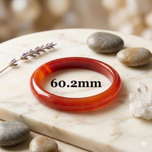 May include: A smooth, polished, translucent red carnelian bangle bracelet with a circular shape. The interior diameter of the bangle is clearly marked as 60.2mm. The bracelet features a vibrant, deep orange-red hue with subtle color variations.