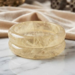 May include: Two stacked bangle bracelets crafted from translucent, pale yellow stone. The material features a natural, fibrous, and crystalline internal texture with a smooth, polished, and rounded exterior surface that reflects light.