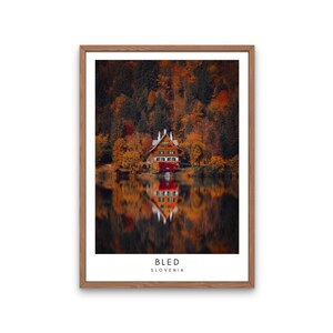 Può includere: Una stampa incorniciata con un edificio rosso e marrone su un lago, circondato da fogliame autunnale. L'immagine si riflette nell'acqua. Il testo "BLED SLOVENIA" è in basso nella stampa.