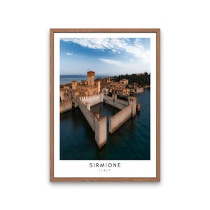 Può includere: Stampa incorniciata di Sirmione, Italia, con un castello medievale su un lago. L'immagine mostra le mura e le torri in pietra del castello, con la scritta "SIRMIONE ITALY" in basso. La cornice è in legno.
