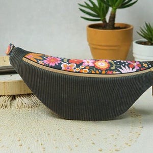 Puede incluir: Un bolso de cintura de pana negro con un estampado floral en la parte superior. La cremallera es dorada. El bolso está sobre una superficie color crema, con plantas en macetas en el fondo.