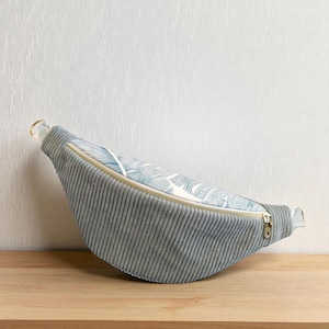 May include: A crescent-shaped fanny pack featuring a grey corduroy body and a top panel with a white and blue wavy line pattern. The bag includes a gold-toned metal zipper closure along the top edge and a small gold-toned metal D-ring on the side.