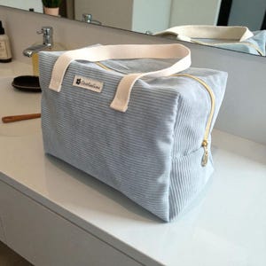 Könnte beinhalten: Hellblaue Cord-Tragetasche mit cremefarbenem Griff und goldenem Reißverschluss. Die Tasche hat eine rechteckige Form und ein kleines Etikett mit Text und einem Blumenlogo. Die Tasche steht auf einer weißen Oberfläche.