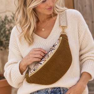 May include: A brown corduroy fanny pack featuring a top panel with a white, brown, and black leaf pattern. The bag includes a gold zipper closure and a wide, adjustable beige strap with a geometric diamond pattern and gold hardware.