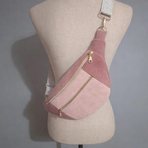 May include: A pink corduroy fanny pack with gold-colored zippers and a gray adjustable strap. The bag is displayed on a mannequin and features a geometric design with varying shades of pink.