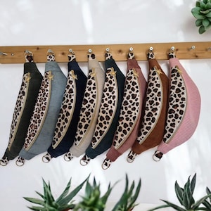 May include: A collection of colorful, corduroy fanny packs hanging on a wooden rack. Each pack features a leopard print accent, a gold zipper, and a matching ring. Colors include green, blue, navy, beige, teal, brown, and pink.