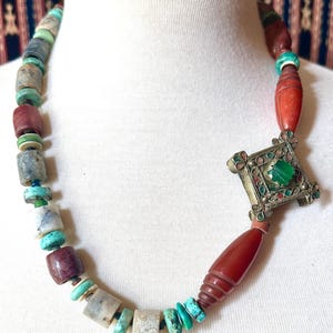 May include: A handcrafted necklace featuring a variety of beads in shades of turquoise, red, and gray. The necklace includes a decorative silver-toned pendant with a green stone center. The beads are of various shapes and sizes.