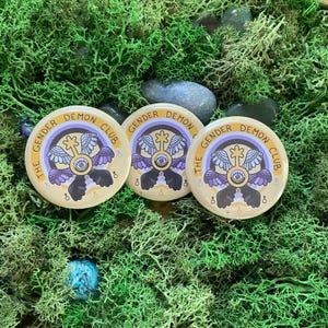 May include: Three round buttons with the text "THE GENDER DEMON CLUB" in a semi-circle. Each button features a central design with wings, an eye, and a symbol. The buttons are beige with purple, black, and yellow accents, set against green moss and stones.