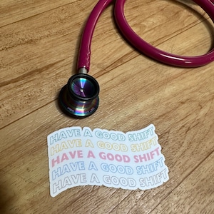 May include: A magenta stethoscope with a rainbow-colored chest piece rests on a wooden surface. A sticker with the text "HAVE A GOOD SHIFT" in multiple colors is also present. Medical equipment.