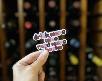 May include: A sticker with the text "drink more wine read more books" in a white and maroon color scheme. The sticker is held in front of a wine rack filled with bottles.