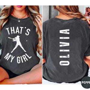 May include: A dark gray t-shirt with a white graphic on the front that says "That's My Girl" and a silhouette of a baseball player. The back of the shirt has the word "Olivia" written vertically in white.