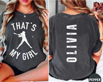 May include: A dark gray t-shirt with a white graphic on the front that says "That's My Girl" and a silhouette of a baseball player. The back of the shirt has the word "Olivia" written vertically in white.