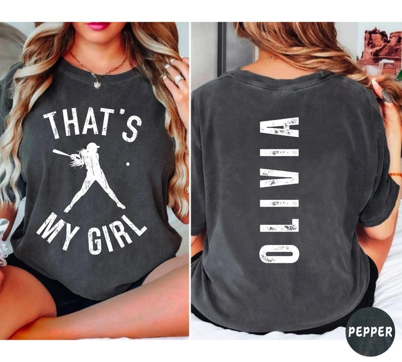 May include: A dark gray t-shirt with a white graphic on the front that says "That's My Girl" and a silhouette of a baseball player. The back of the shirt has the word "Olivia" written vertically in white.