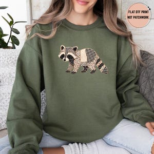 May include: An olive green crewneck sweatshirt featuring a graphic of a raccoon composed of various patterned segments including gingham, leopard print, and solid beige. A circular text overlay in the top right corner reads FLAT DTF PRINT NOT PATCHWORK.