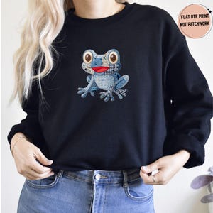 May include: A black crewneck sweatshirt featuring a central graphic of a frog with a blue and white patchwork-style pattern and a bright red mouth. A circular badge in the top right corner contains the text: FLAT DTF PRINT NOT PATCHWORK.