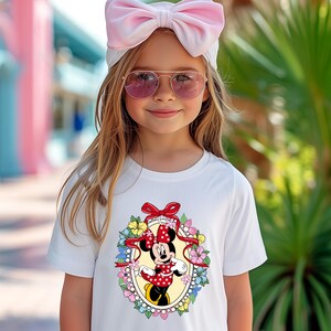 Peut inclure: Une jeune personne portant un t-shirt blanc avec un motif de Minnie Mouse dans un cadre ovale avec des fleurs. La personne porte &eacute;galement un bandeau rose avec un n&oelig;ud et des lunettes de soleil roses.