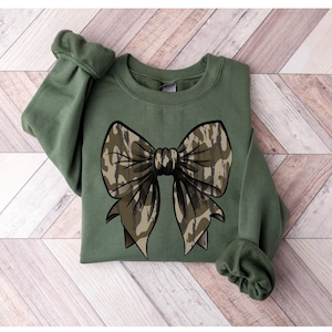May include: A green sweatshirt with a large camouflage bow on the front. The bow is a light brown and green color.