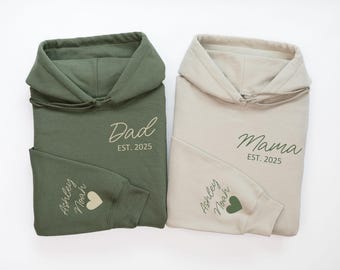 May include: Two green and beige hooded sweatshirts with the words "Dad Est. 2025" and "Mama Est. 2025" printed on the front. The sleeves have the names "Ashley Noah" and a green heart printed on them.