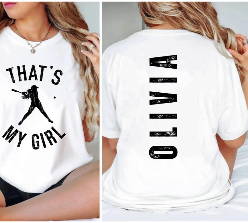 May include: White t-shirt with a black graphic of a baseball player and the text "That's My Girl". The back of the shirt has the name "Olivia" printed vertically in black.
