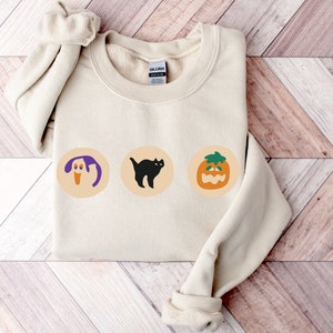 May include: A cream-colored sweatshirt featuring three Halloween-themed graphics. The graphics include a ghost, a black cat, and a jack-o'-lantern, each set within a circular design. The sweatshirt is folded, with the sleeves visible.